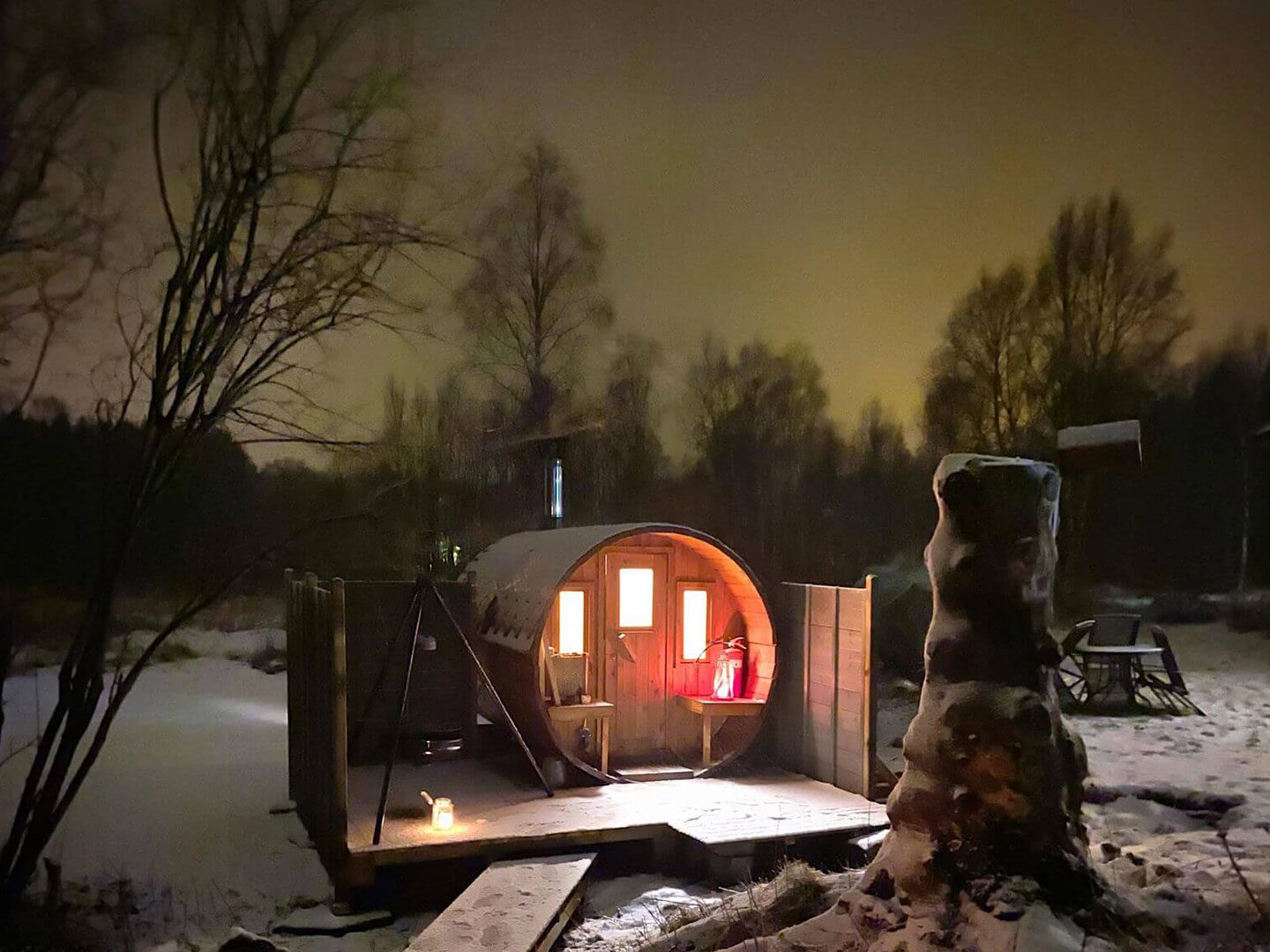 A picture taken outside in the norwegian wilderness with snow and a round wooden cylinder sauna lit up from the inside
