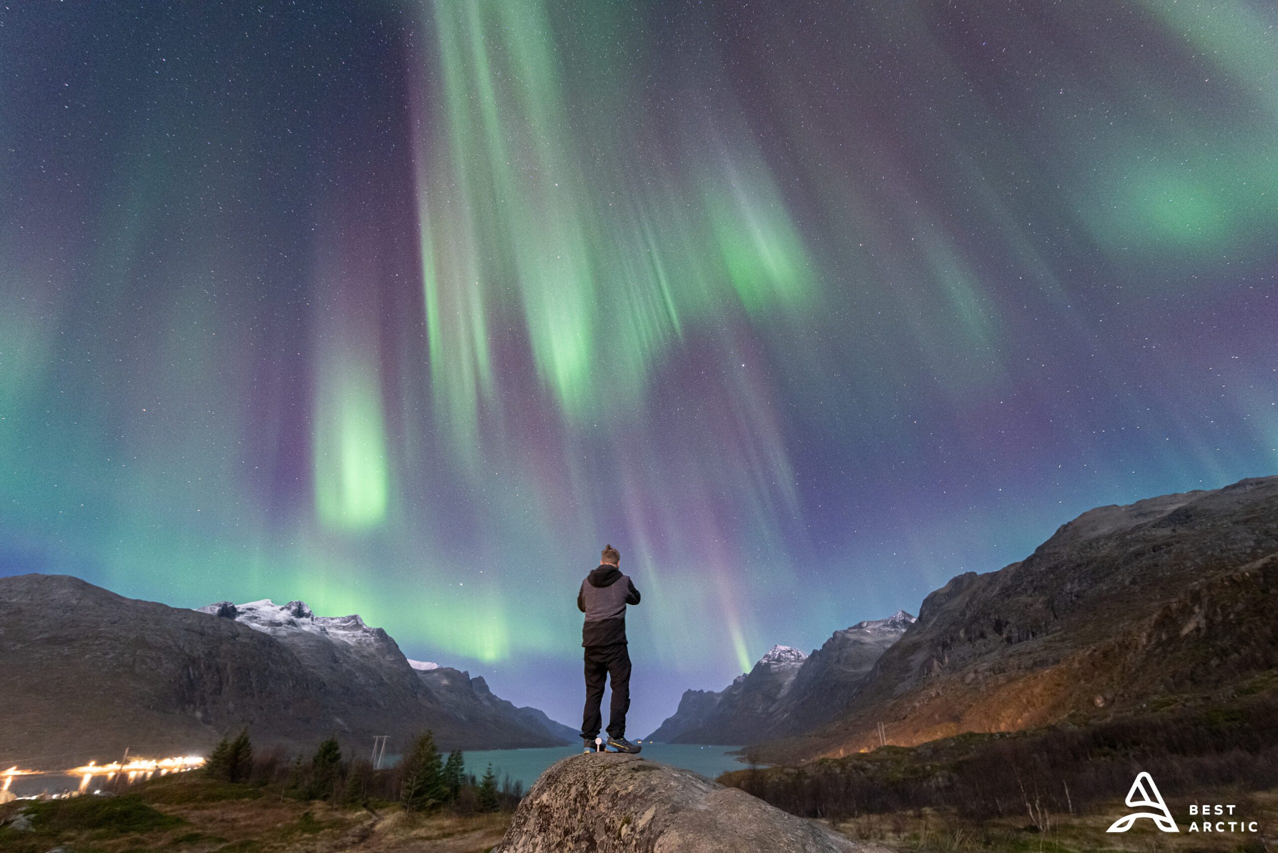 A picture taken from the back of a person witnessing a colorful aurora borealis surrounded by a magnificent fjord