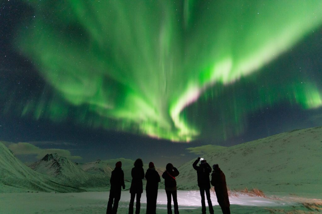 A group of people watching the Northern Lights in a area with high snow covered mountains