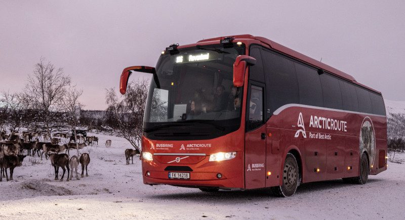 The Arctic Route bus surrounded by snow. On the left of the image, there are reindeers.