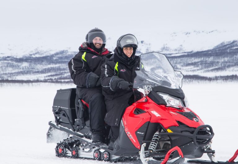 A man and a woman on a red snowmobile wearing winter thermal suits and helmets surrounded by snowy mountains and forests