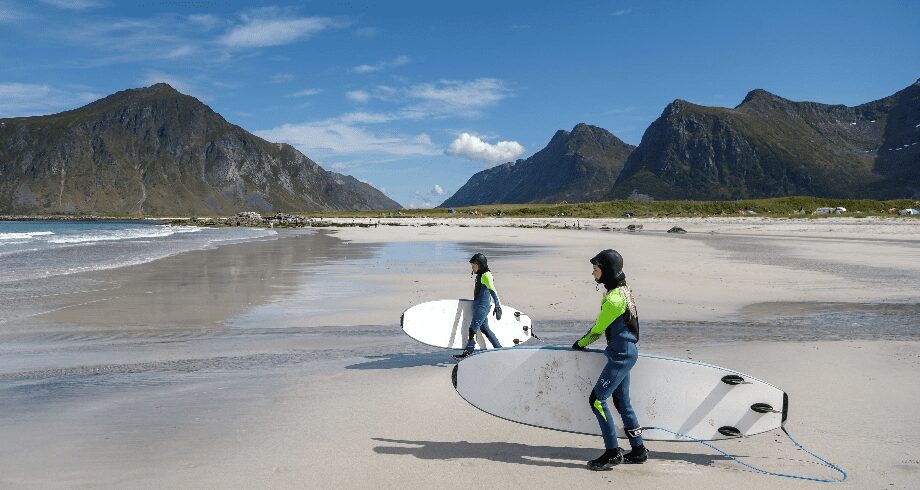 2 people surfing on a white sand beach surrounded by green mountains