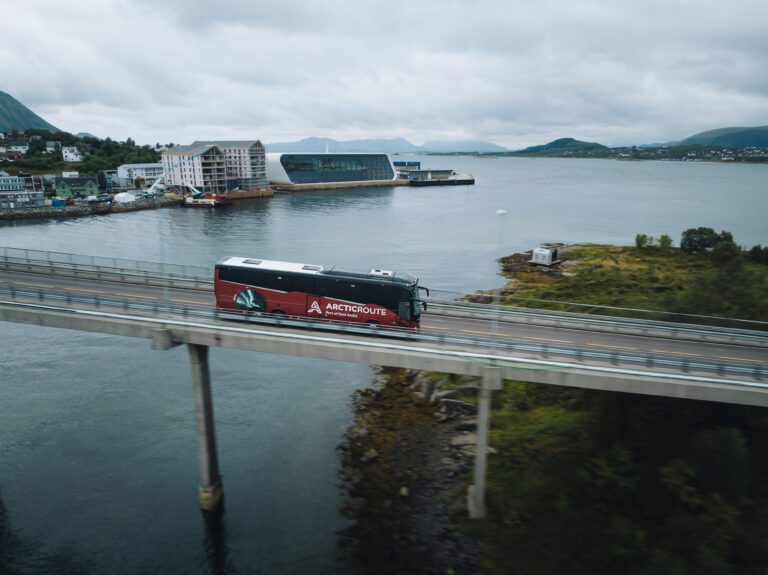 A red bus crossing a body of water with buildings in the background.