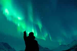Person pointing at vivid green northern lights in admiration, and surrounded by snowy mountains