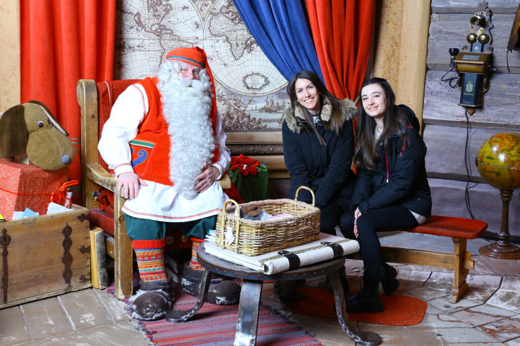 Two women sitting next to Santa Claus, with a large world map, red and blue velvet curtains on the background, and a basket on a table in front of them.