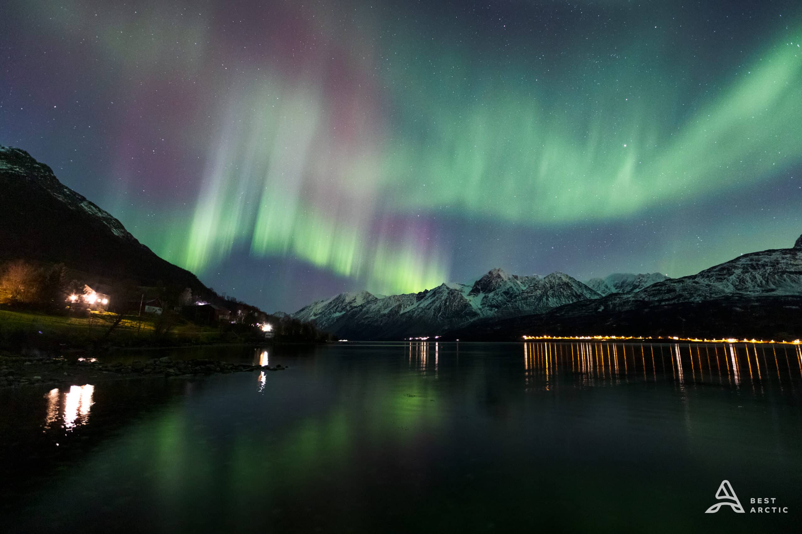 Snowy mountains reflected on calm water at night, with street lights along the shoreline and green and purple northern lights with stars in the sky.