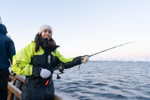 Young woman in a thermal suit fishing from the wooden boat Hermes II in Arctic waters with snowy mountains in the background.