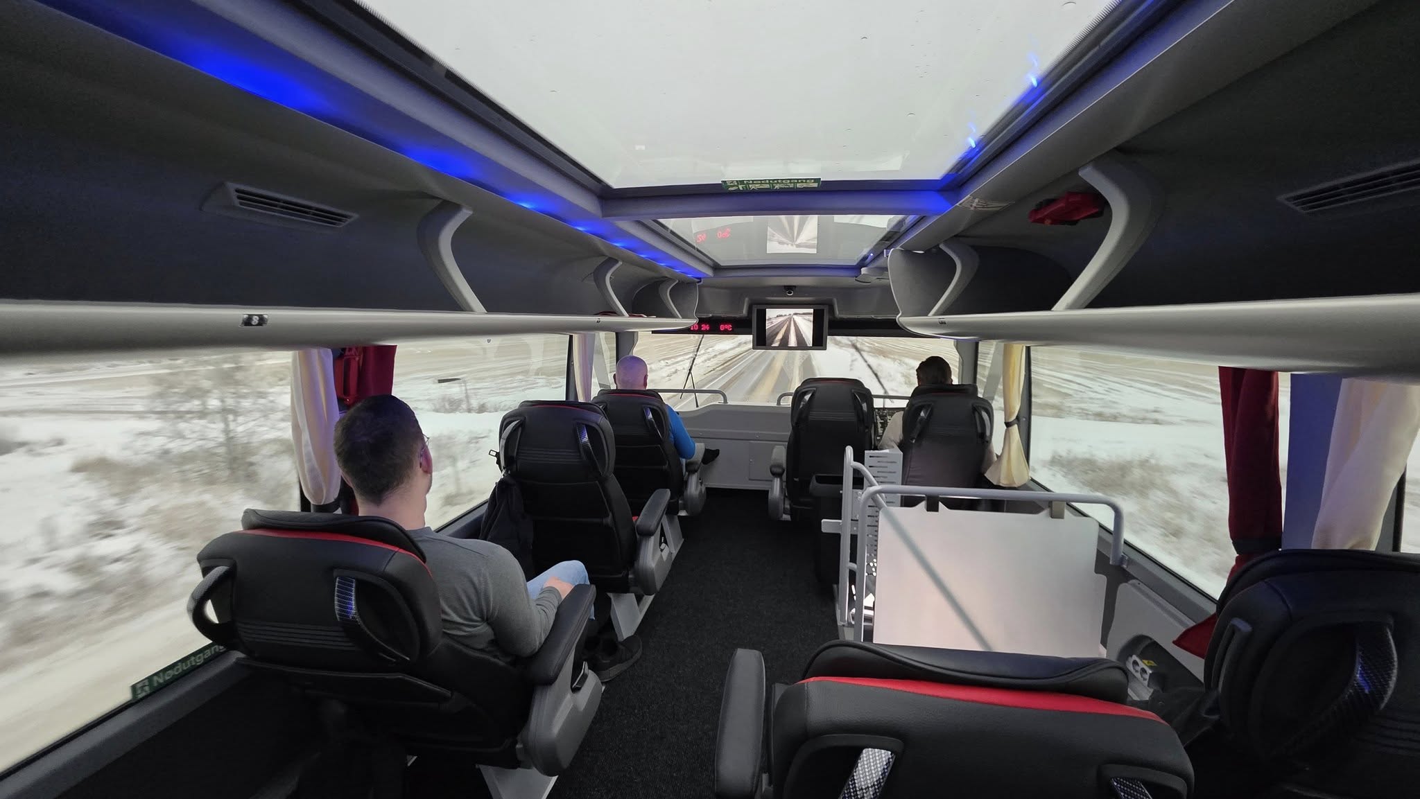 Interior of the Aurora Express bus top deck with passengers seated, panoramic windows, skylight panels, and snowy landscape visible outside.