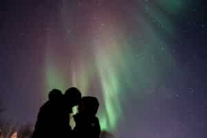 Silhouette of a couple touching foreheads beneath green and purple northern lights and a star-filled night sky.