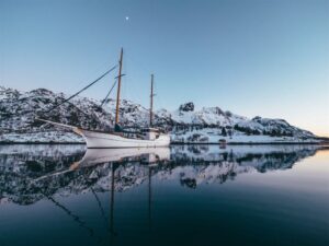 White wooden traditional sailing boat in a calm fjord surrounded by snow-covered mountains