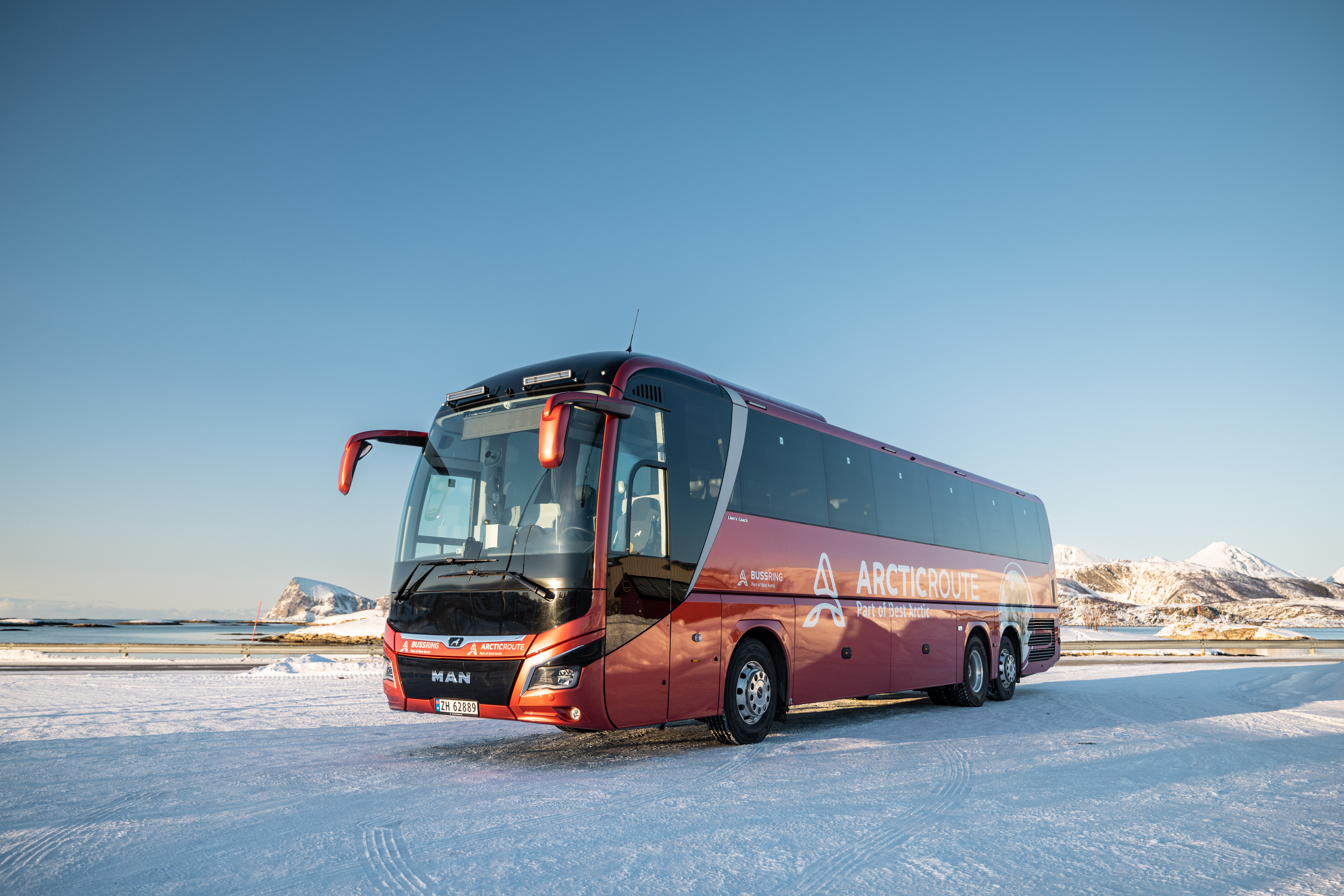 Red Arctic Route bus from Best Arctic parked by a beach with snowy mountains in the background in northern Norway
