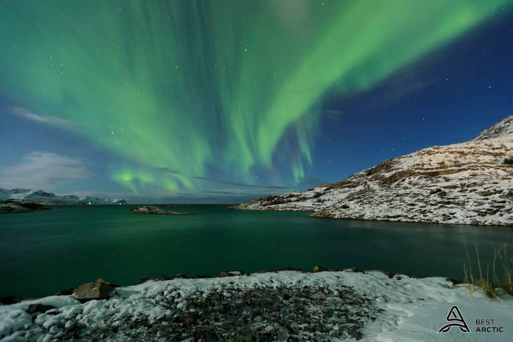 Snowy mountains in the Tromsø region overlooking the Arctic sea, under green Northern Lights in a starry sky