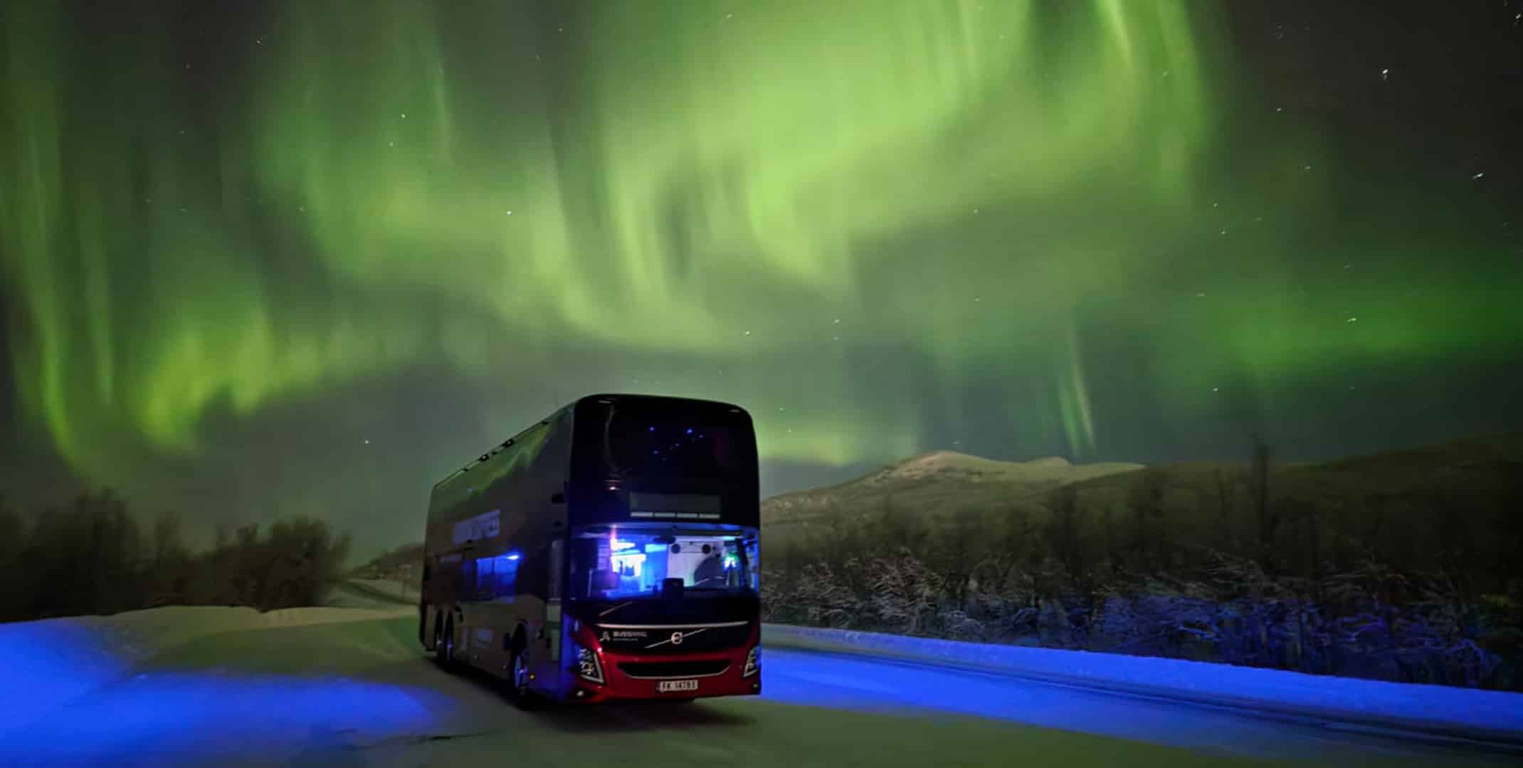 Red Aurora Express double-decker bus driving on a snowy road under vivid green northern lights with mountains and a clear night sky