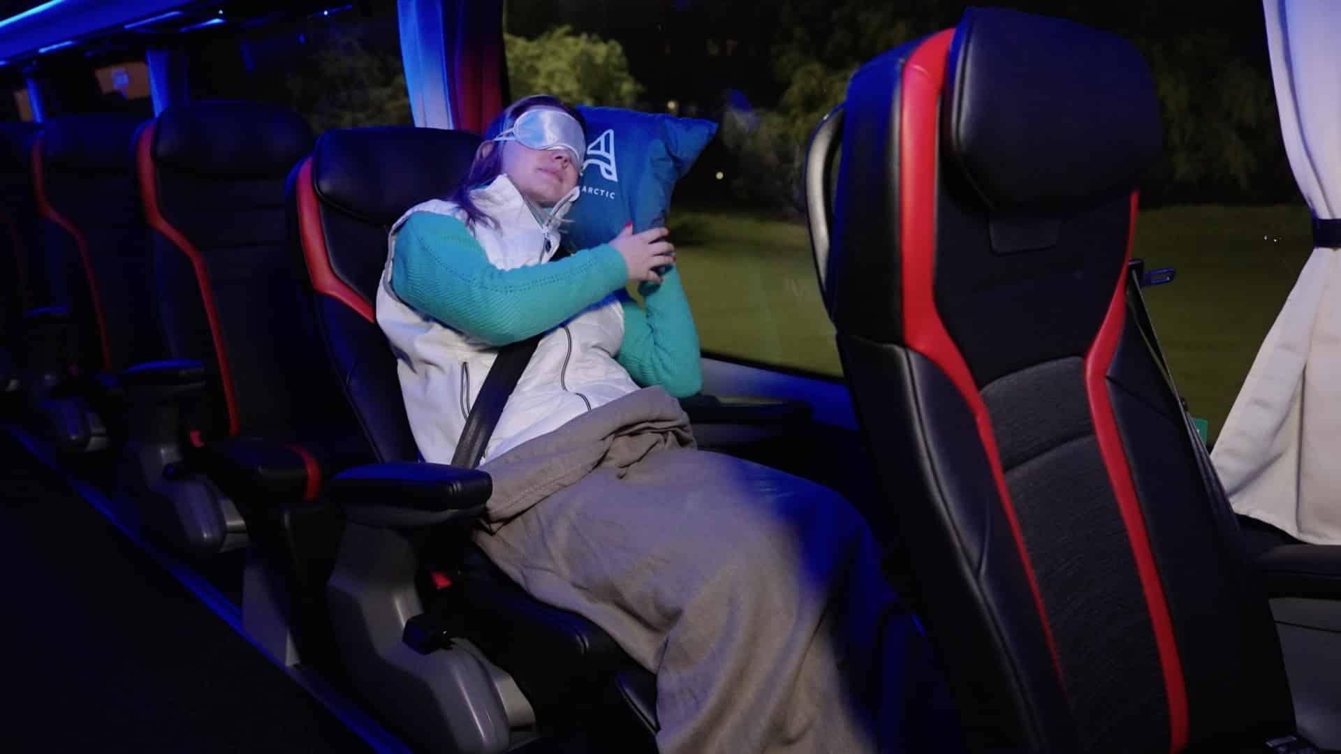 A woman wearing an eye mask is sleeping on the Aurora Express bus, leaning against the window with a pillow.